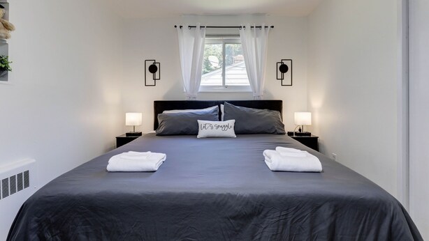 A neatly made bed with a dark gray bedspread, two pillows, and a decorative pillow with the text "It's snuggle." There are two bedside tables with lamps, a window with white curtains, and a wall-mounted light fixture. - Modern living for visit DC/UMD, 2 bedrooms, 4 beds with central AC/heating (Hyattsville)