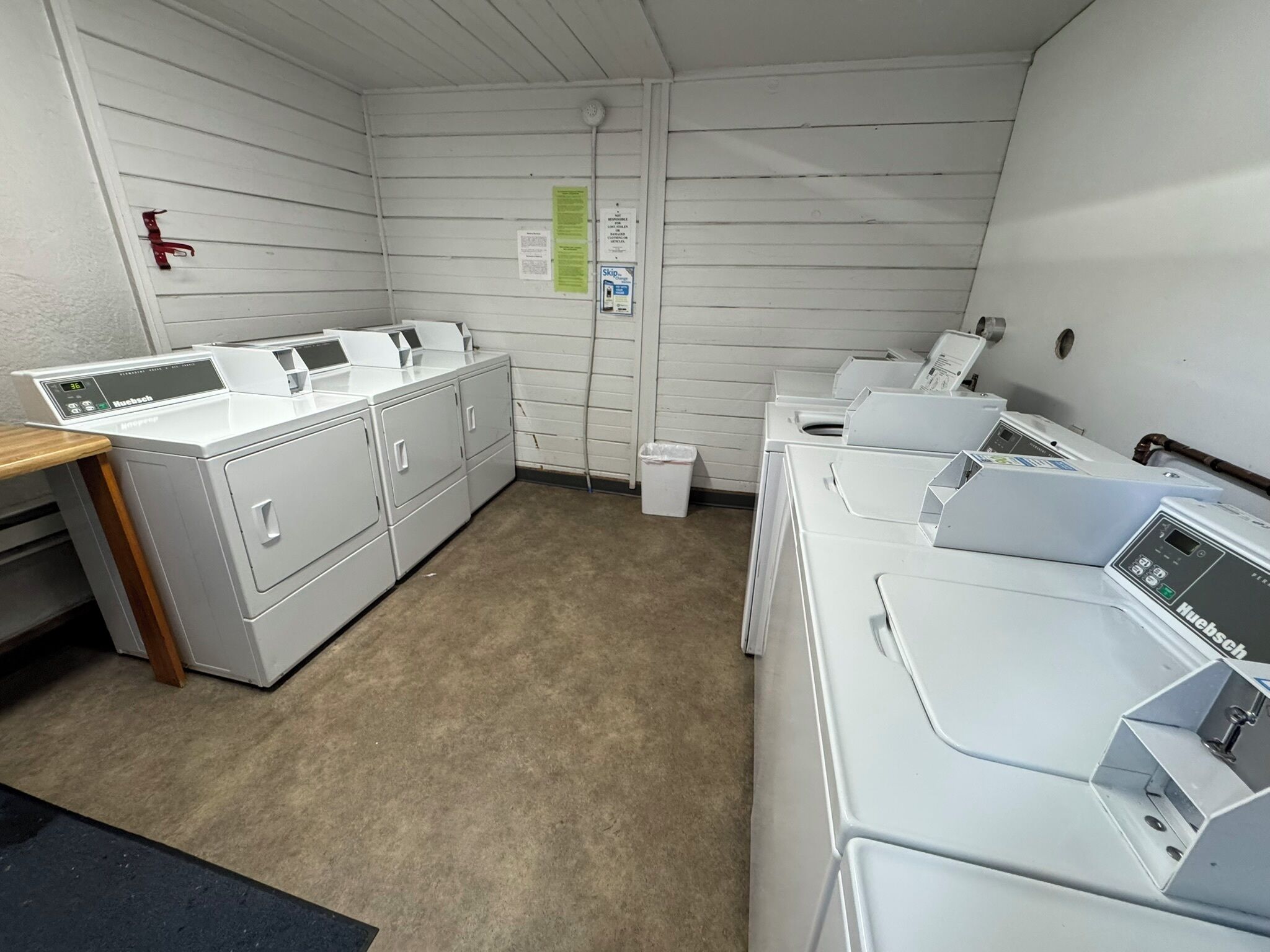 Une buanderie équipée de plusieurs machines à laver et de plusieurs sèche-linge, avec une table en bois et une poubelle.