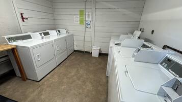 Une buanderie équipée de plusieurs machines à laver et de plusieurs sèche-linge, avec une table en bois et une poubelle.