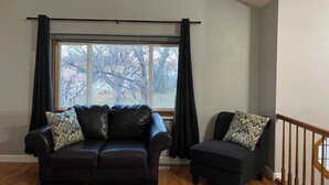 A living room with a black leather sofa and a matching armchair, both with patterned cushions. A large window with curtains is visible, and a wooden staircase is on the right. - Millard West Omaha 4 bed, 3 bath (Omaha)