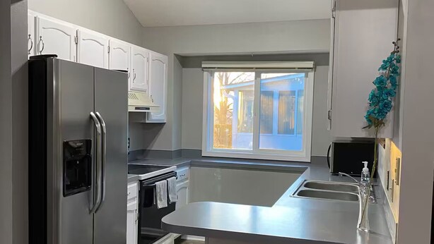 A modern kitchen with white cabinets, a stainless steel refrigerator, an oven, a sink, and a window with a view of the outside. - Millard West Omaha 4 bed, 3 bath (Omaha)