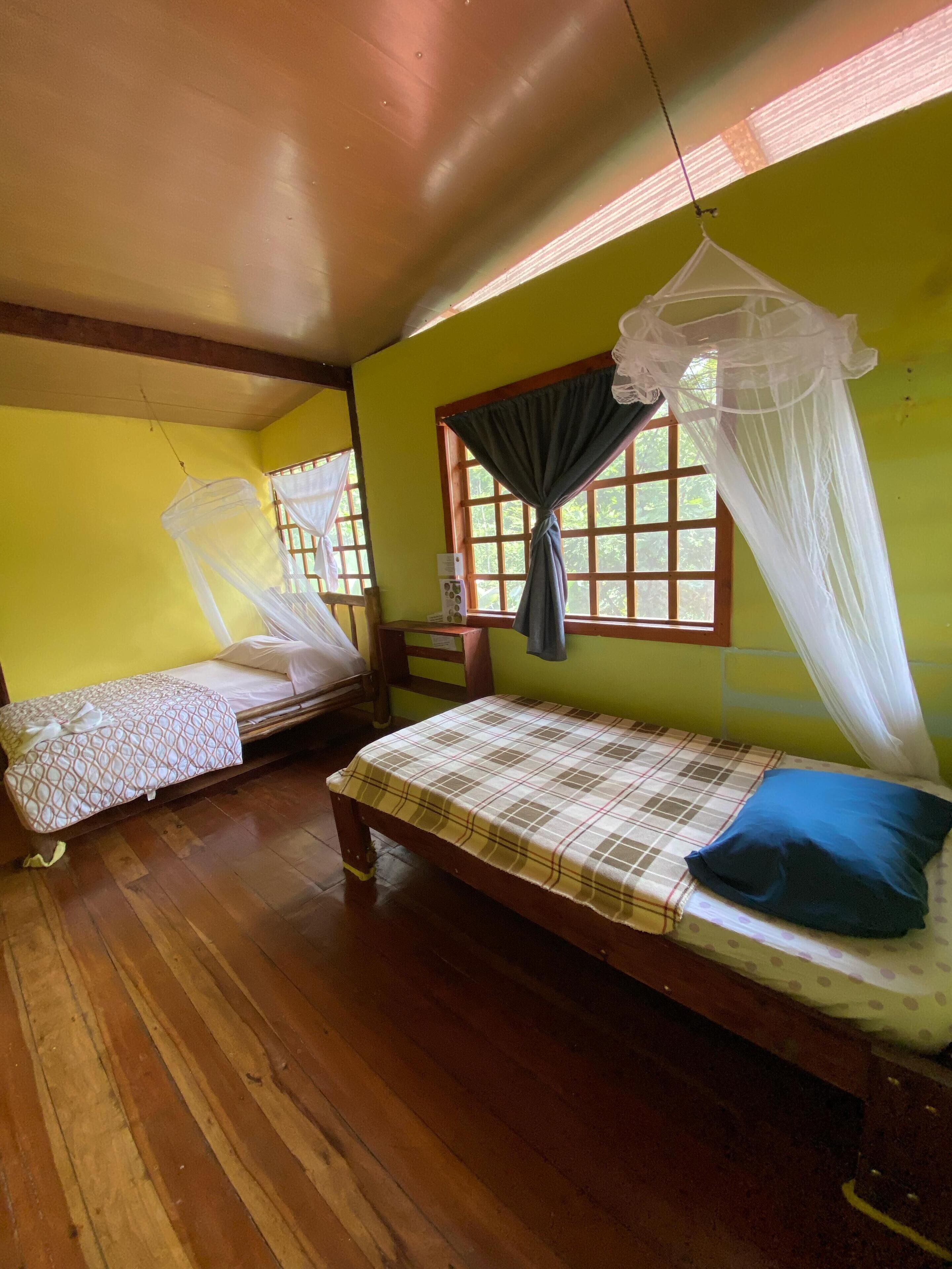 A room with two wooden beds, one with a canopy and one without. There is a window with a curtain and a mosquito net hanging above the bed.