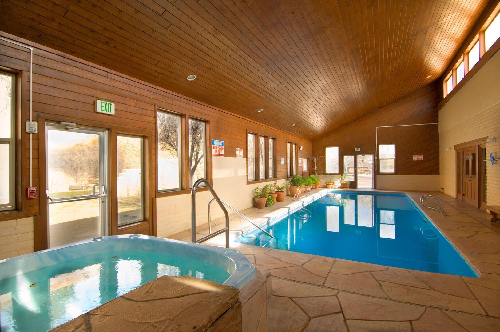 Una piscina interior con techo de madera y suelo de piedra, rodeada de ventanas y plantas en macetas.