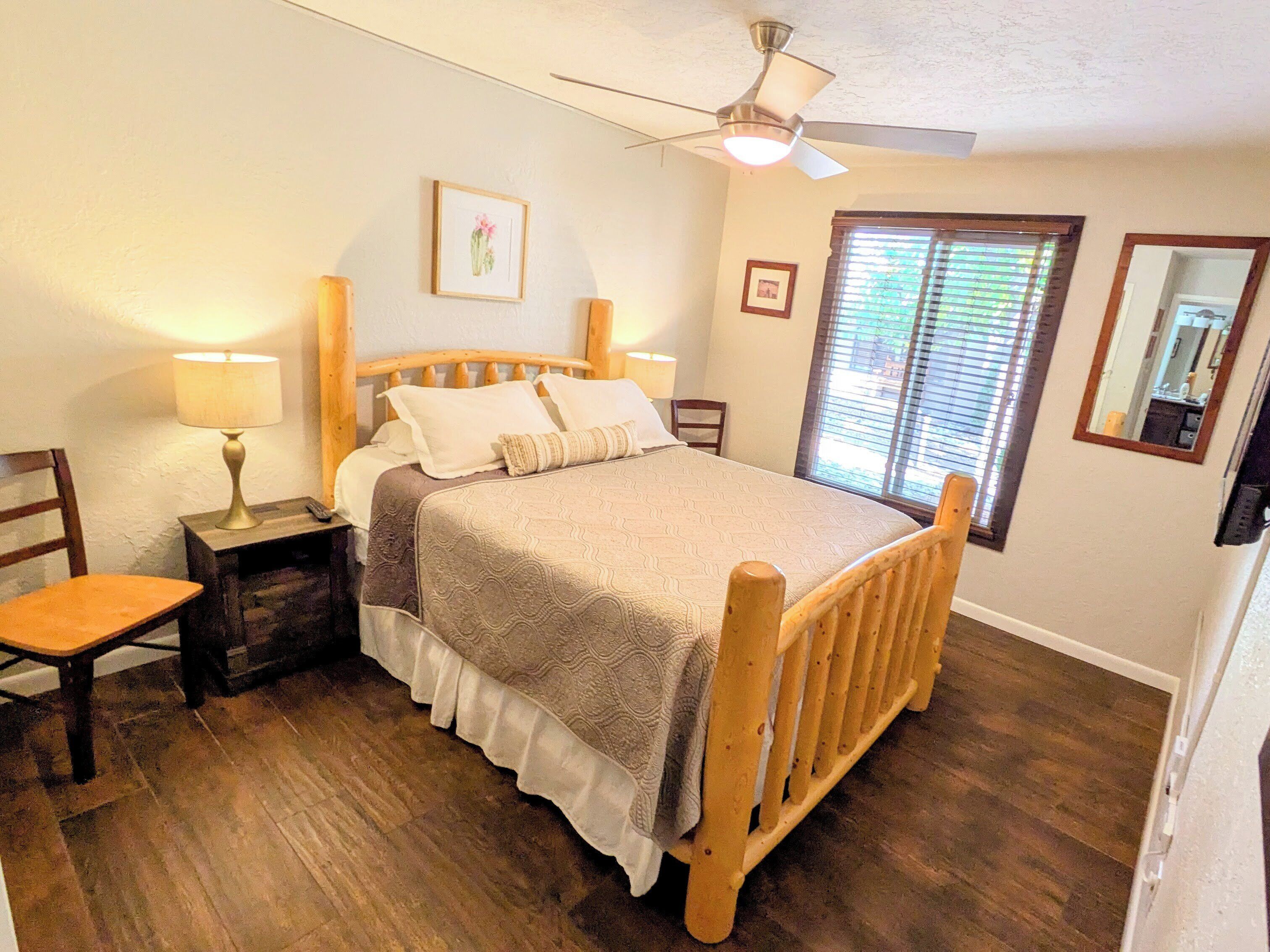 A bedroom with a wooden bed, two bedside tables with lamps, a ceiling fan, a window with blinds, and a framed picture on the wall.