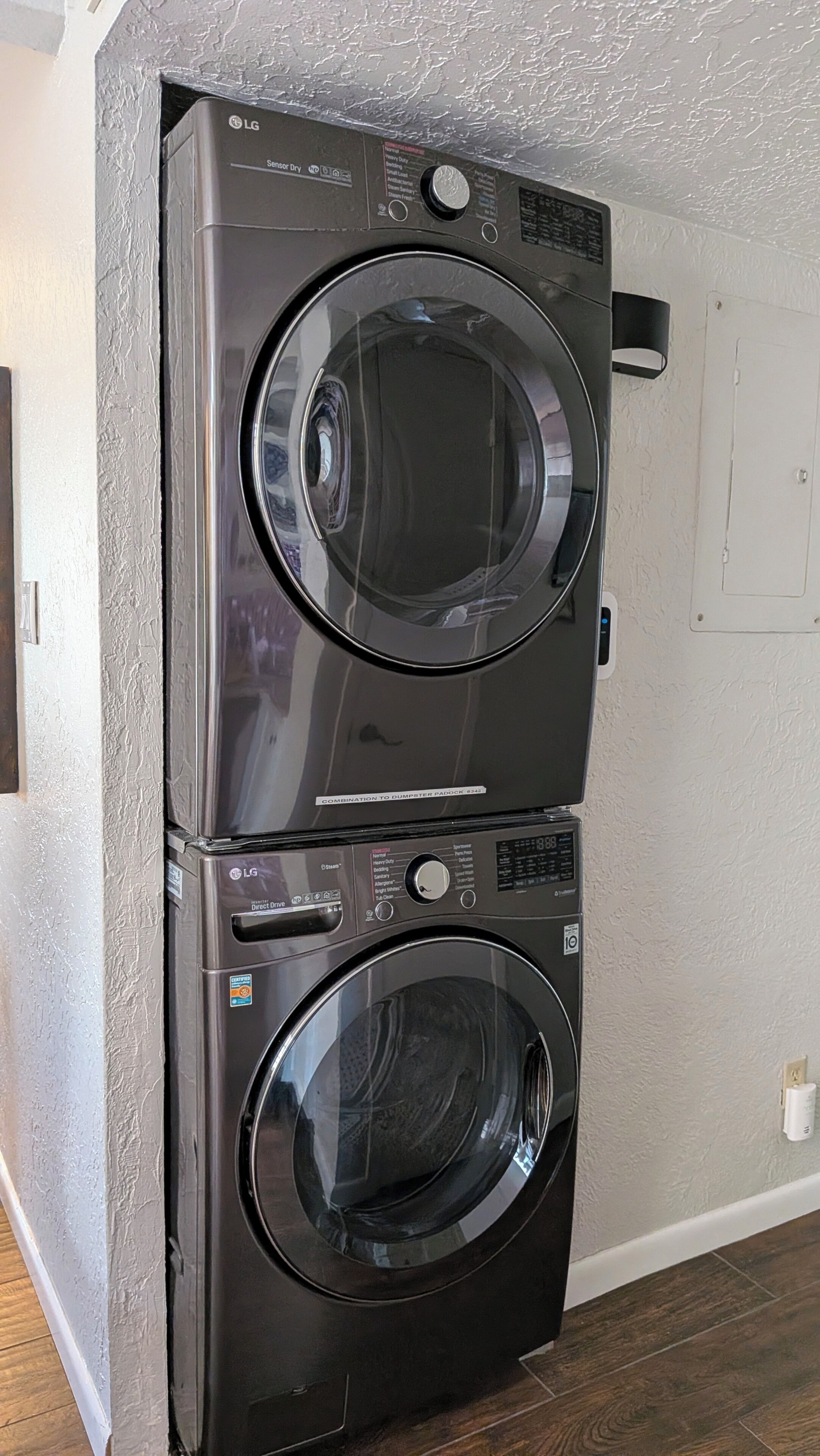 Un appareil combiné lave-linge et sèche-linge, avec un panneau de commande, installé dans une pièce dont les murs sont recouverts d’un revêtement texturé et dont le sol est en bois.