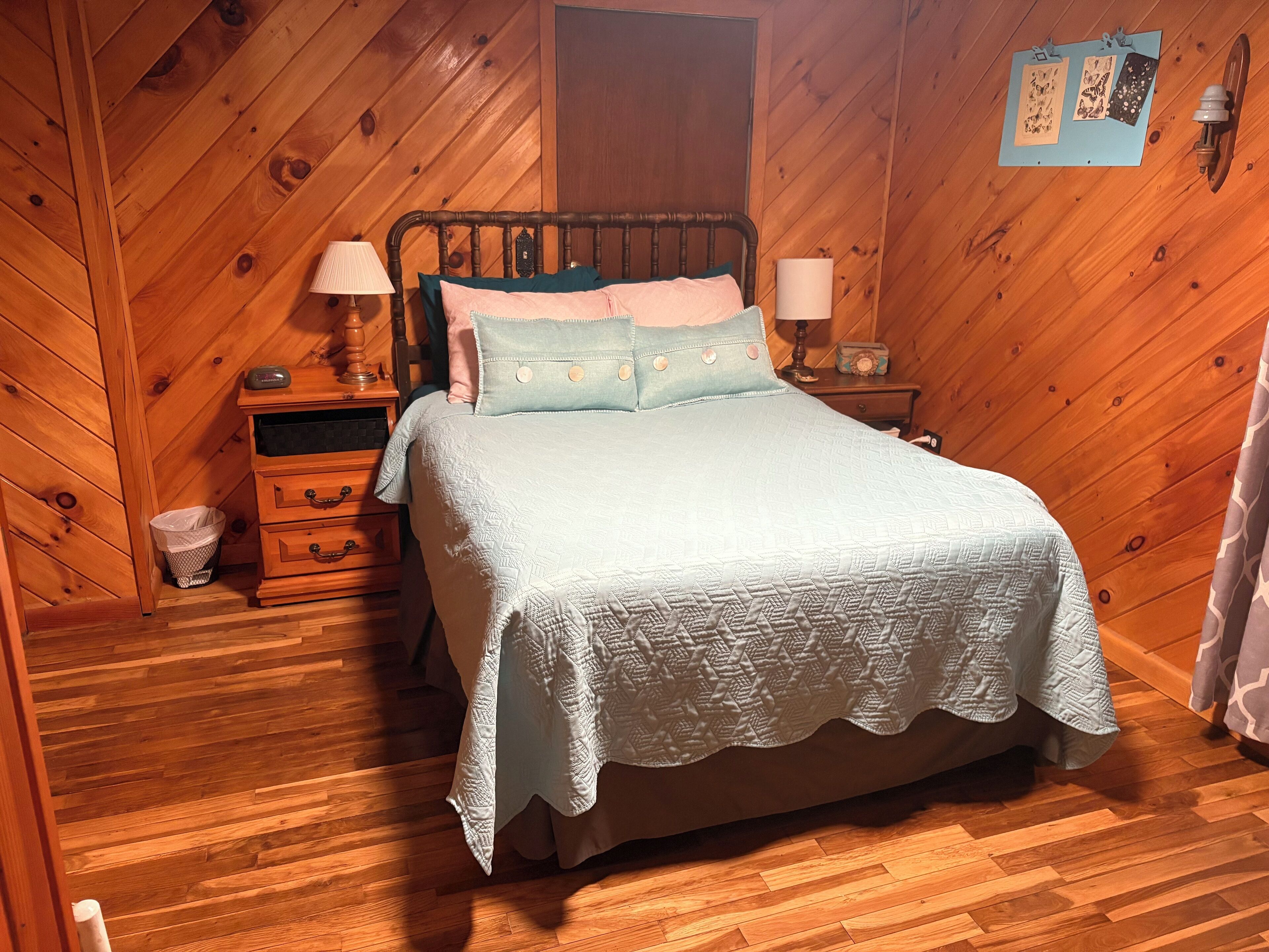 A cozy bedroom with a wooden sloped ceiling, a bed with a quilted cover, two bedside tables with lamps, and a wall-mounted bulletin board.