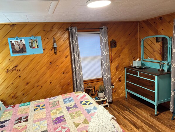 Un dormitorio acogedor con paredes de madera, una cama con colcha colorida, un aparador con espejo y una ventana con cortinas.