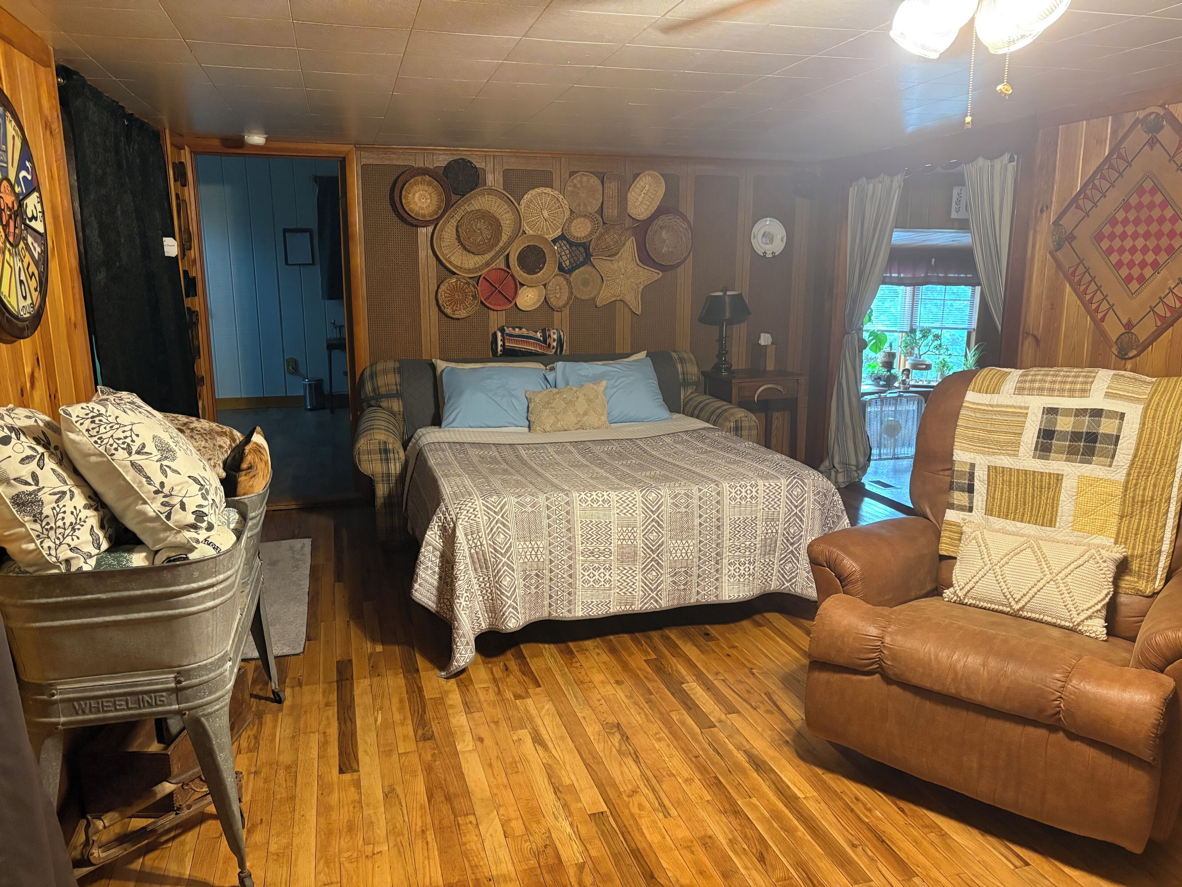 A cozy room with a wooden floor, a bed with patterned bedding, a brown leather armchair, and a vintage wheelbarrow.