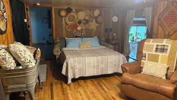 A cozy room with a wooden floor, a bed with patterned bedding, a brown leather armchair, and a vintage wheelbarrow.