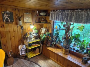 Een houten interieur met een plank waarop verschillende spullen staan, een raam met planten en een lamp.
