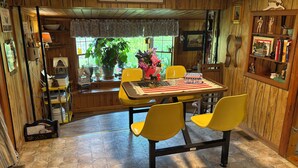 Een houten interieur met een 'Welcome'-bord, een eettafel met gele stoelen, een raam met planten en een boekenkast met boeken en beeldjes.