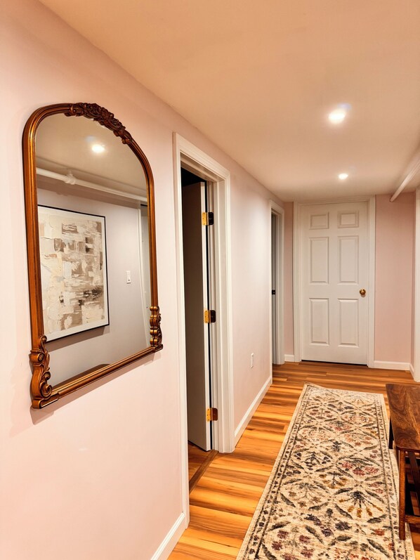 A hallway with a wooden floor, a patterned rug, a large mirror, and a door leading to another room.