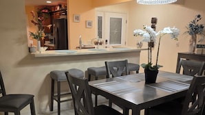 A dining area with a rectangular table, six chairs, and a potted orchid. A kitchen is visible in the background with a refrigerator and a sink.