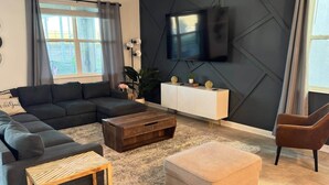 Ein modernes Wohnzimmer mit einer Eckcouch, einem Holztisch, einem beige Ottomanen, einem an der Wand montierten Fernseher und einem weißen Beistelltisch mit Dekorationselementen.