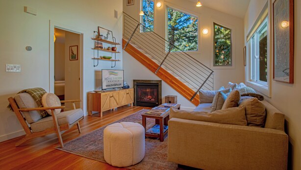 A spacious living room with a wooden staircase, a fireplace, and large windows offering views of trees. - 2BD, 2BA Ketchum Condo – Newly Remodeled & Central (Ketchum)