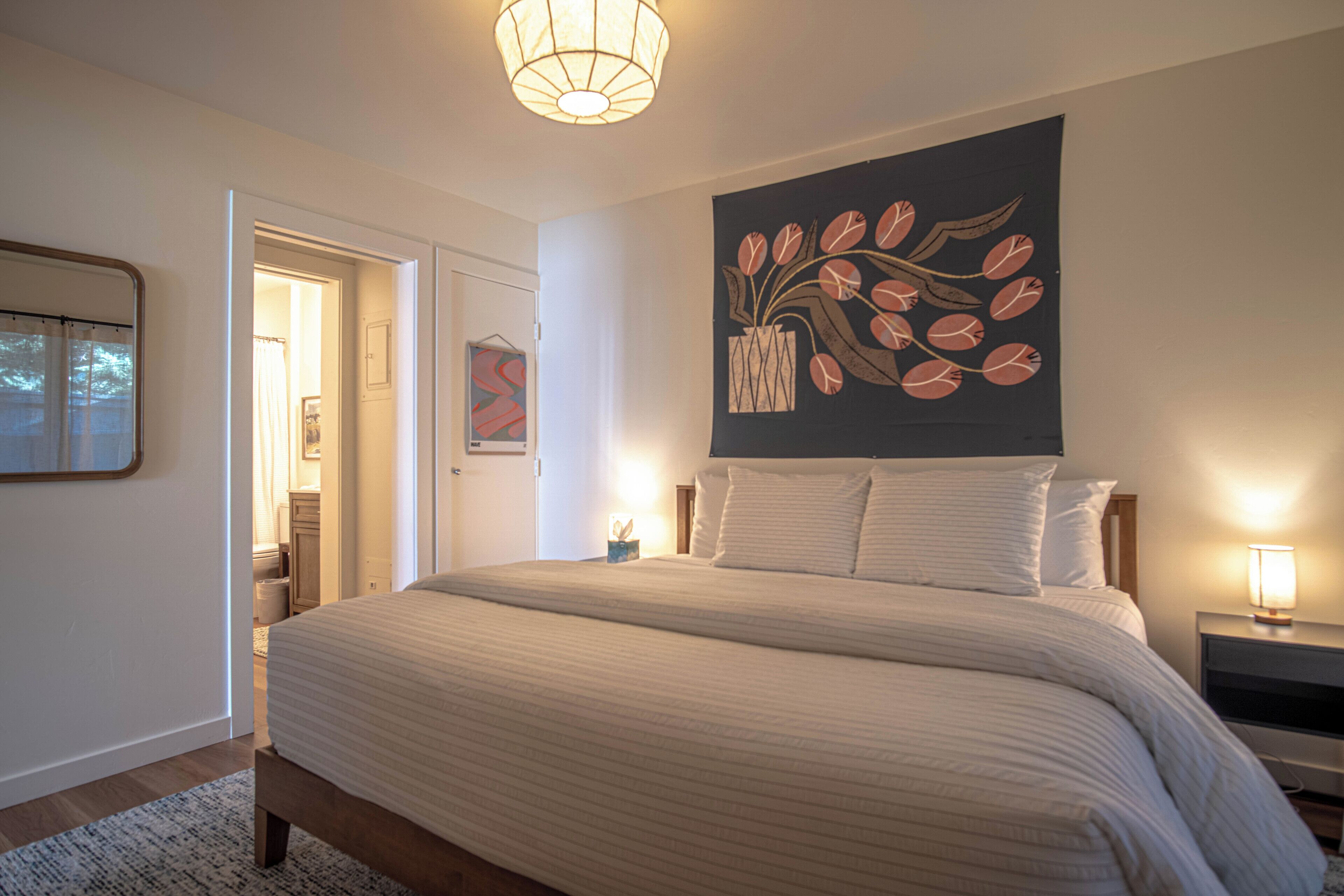 Una camera da letto con un letto grande, un comodino con una lampada, una porta che dà su un'altra stanza e una parete decorata con un dipinto floreale.