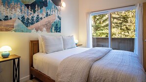 A bedroom with a large bed, a nightstand with a lamp, a wall painting, and a window with a view of trees. - 2BD, 2BA Ketchum Condo – Newly Remodeled & Central (Ketchum)