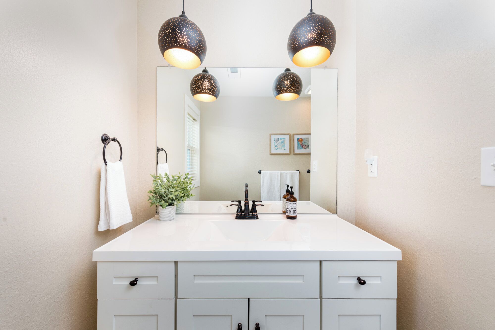 A bathroom with a white vanity, three pendant lights, a mirror, a towel rack, a potted plant, and a soap dispenser.