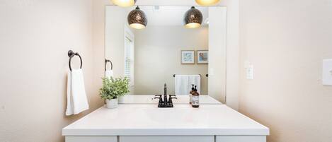 A bathroom with a white vanity, three pendant lights, a mirror, a towel rack, a potted plant, and a soap dispenser.