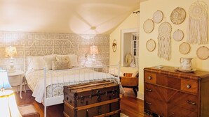 Una camera da letto con una testiera decorata, un settimino in legno, un baule vintage e degli acchiappasogni appesi al muro.