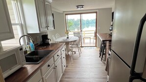 A kitchen with white cabinets, a microwave, a sink, and a refrigerator. There is a dining table with chairs and a view of a lake through a large window. - Beautiful Lakefront Escape with Private Dock & Wildlife Views (Eau Galle)