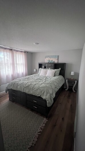 Un dormitorio con cama, mesita de noche, lámpara, alfombra y ventana con cortinas.