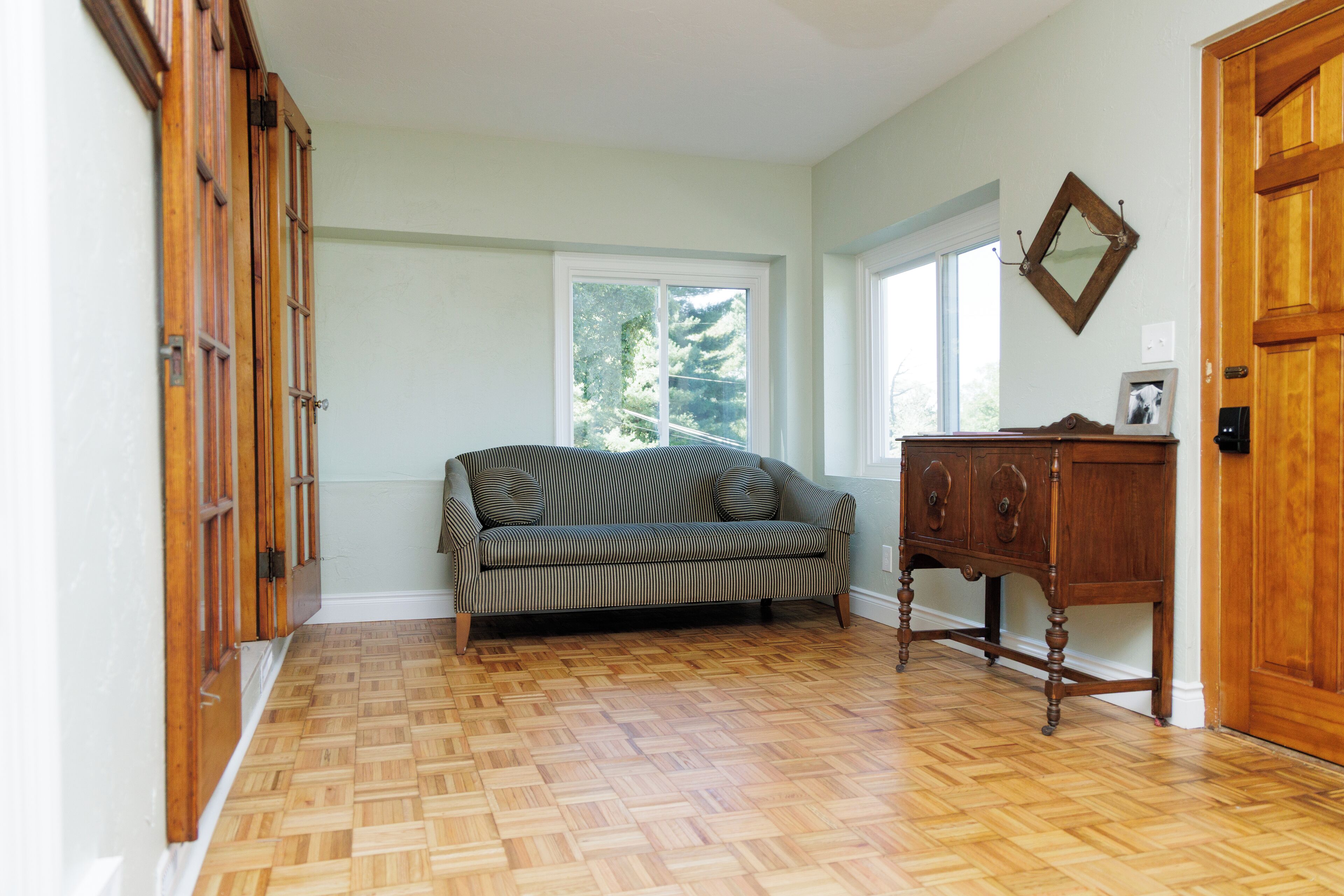 A room with a herringbone-patterned wooden floor, a striped sofa, and a wooden cabinet with a mirror and picture frame.