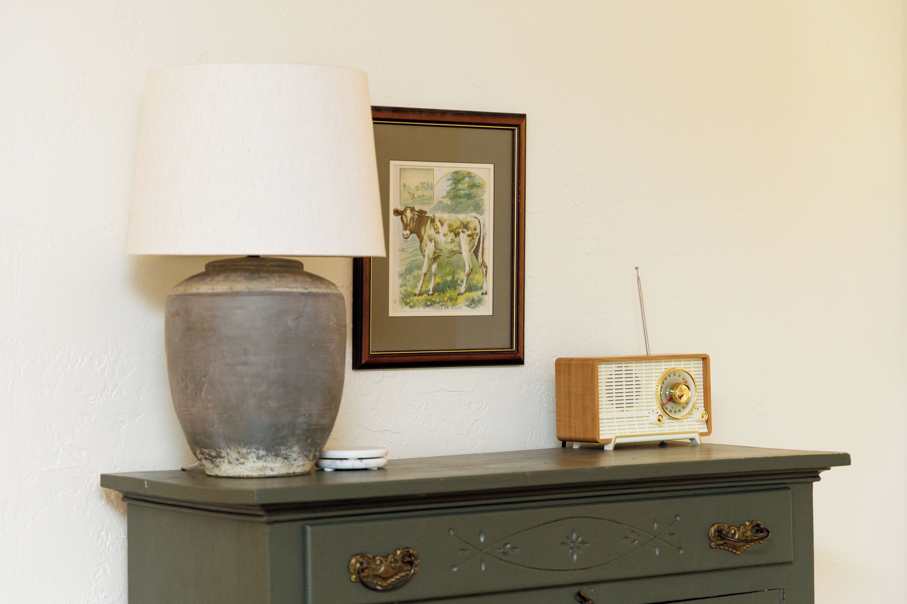 Une vieille radio posée sur une commode, une lampe et un tableau représentant une vache accroché au mur.