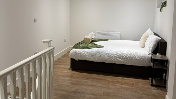 A bedroom with a bed, a nightstand, a white railing, and a window with blinds.
