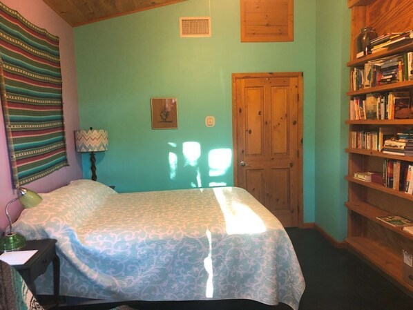 A bedroom with a bed, a nightstand, a lamp, a bookshelf, a door, and a window.
