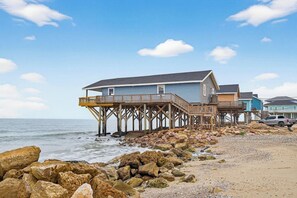 A coastal property with a wooden pier extending into the sea, a beach with rocks, and a clear sky.