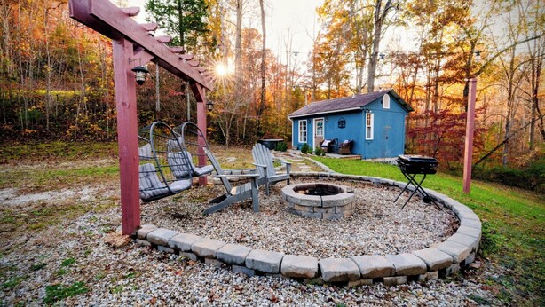 Un patio trasero con un brasero, sillas Adirondack, una pérgola y una casa pequeña de color azul.
