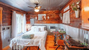 Una habitación rústica de madera con cama, ventilador de techo, una pequeña mesa con tocadiscos y una ventana con cortinas.