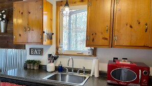 Una cocina con armarios de madera, un lavamanos de acero inoxidable, un microondas rojo y una ventana con vista a árboles.