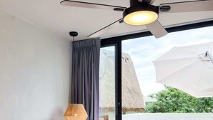 Une chambre d’hôtel avec un lit, un ventilateur de plafond, une fenêtre avec des rideaux et une vue sur une construction au toit de chaume et sur la végétation environnante.