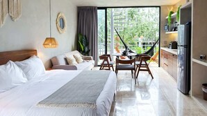 Une chambre d’hôtel moderne dotée d’un grand lit, d’un coin repas avec une table et des chaises, d’un coin cuisine avec un réfrigérateur et d’un balcon offrant une vue sur la verdure.