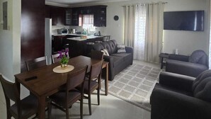 A modern living room with a dining area, a kitchen in the background, and a flat-screen TV mounted on the wall.