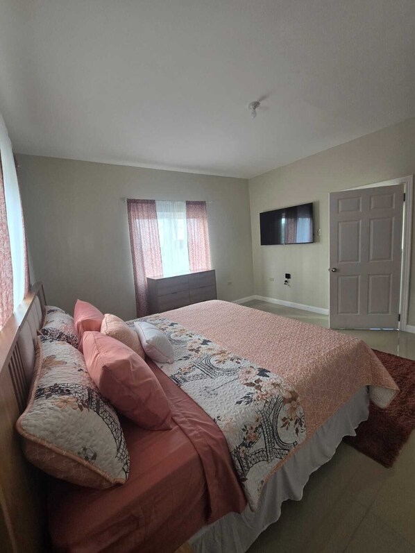 A bedroom with a bed, a dresser, a TV mounted on the wall, and a door. - A Rejuvenating & Relaxing Retreat overlooking Ocho Rios. (Ocho Rios)