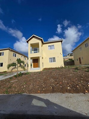 A two-story residential building with a flat roof, a balcony on the upper floor, and a small garden area in front. - A Rejuvenating & Relaxing Retreat overlooking Ocho Rios. (Ocho Rios)