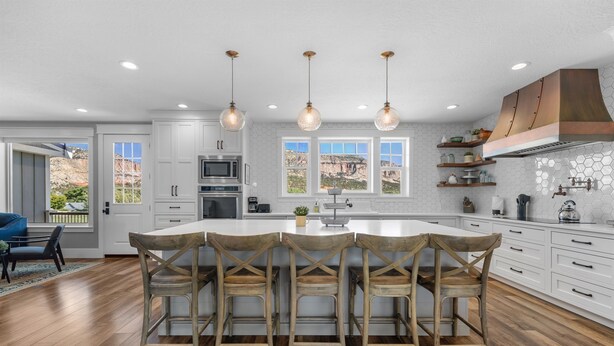 A modern kitchen with white cabinetry, a large island with bar stools, and a view of the mountains through large windows. - East Zion Modern Farmhouse, Sleeps 14, Hot Tub, Fire Pit & Chef Kitchen (Orderville)