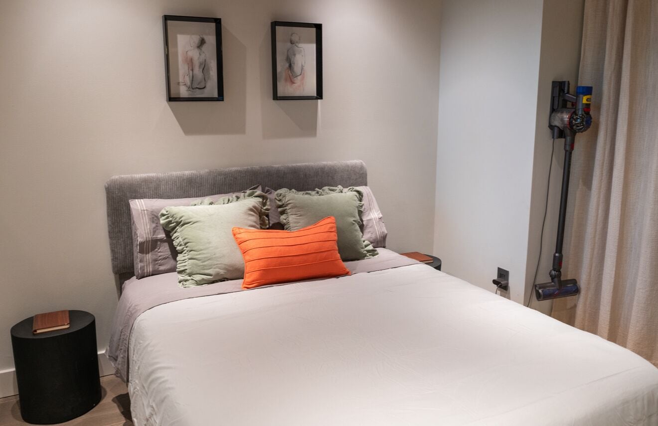 Ein ordentlich bezogenes Bett mit weißer Decke und mehreren Kissen, ein grauer Kopfteil und zwei gerahmte Kunstwerke an der Wand.