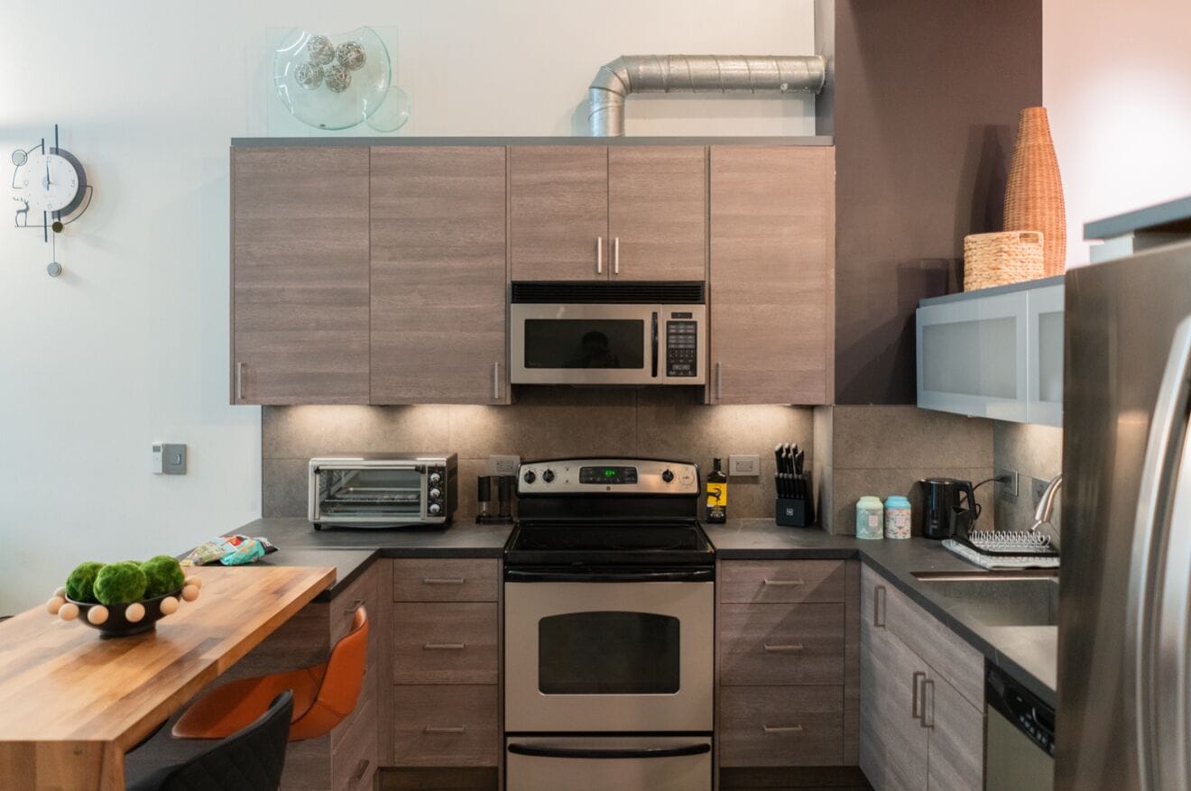 A modern kitchen with a built-in microwave, oven, and toaster. There is a wooden table with a bowl of decorative balls and a chair.