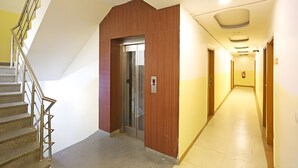Corridoio di un hotel con una scala a sinistra, un ascensore e un lungo corridoio con porte a destra.