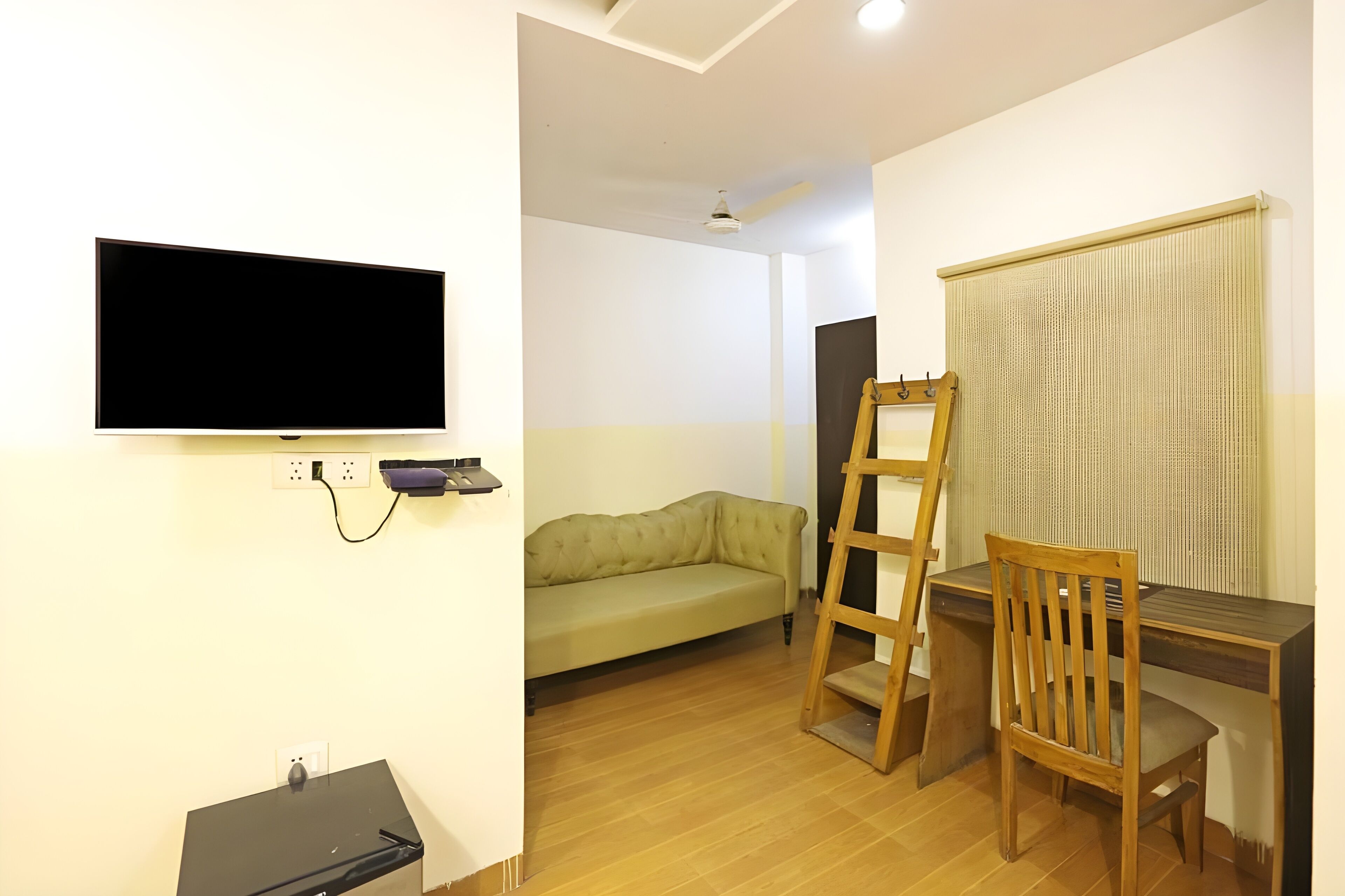 A room with a flat-screen TV mounted on the wall, a wooden desk with a chair, a green sofa, and a ladder leaning against the wall.