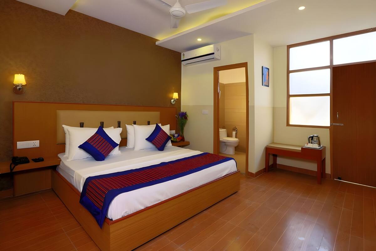 A hotel room with a large bed, bedside tables, a small bench, an air conditioning unit, a ceiling fan, and a view of the outside through a window.