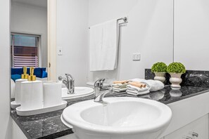 A modern bathroom with a large sink, two faucets, and a mirror. There are towels, toiletries, and two potted plants on the counter. - Modern Victorian Luxury in Melbourne CBD | 3BR 2BA (Melbourne)