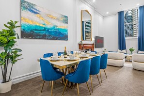 A dining area with a table set for six, blue chairs, a large abstract painting, a mirror, and a view of a building through a window. - Modern Victorian Luxury in Melbourne CBD | 3BR 2BA (Melbourne)