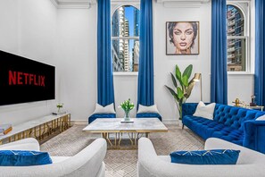 Ein modernes Wohnzimmer mit großem Flachbildfernseher, blauen Vorhängen, einem blauen Sofa und einer Topfpflanze.