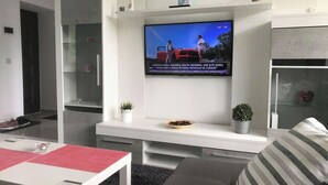 A modern living room with a flat-screen TV mounted on a white wall, a grey sofa, a small table with a red placemat, and a glass cabinet.