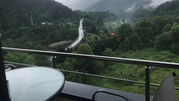 A balcony with a table and chairs overlooking a valley with a road and a bridge.